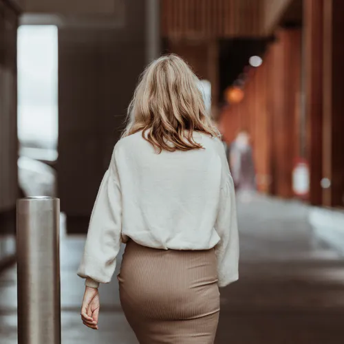 The back view of a hand knit sweater, paired with a dark beige skirt. She has blonde hair and walks down an outdoor, retail alleyway.